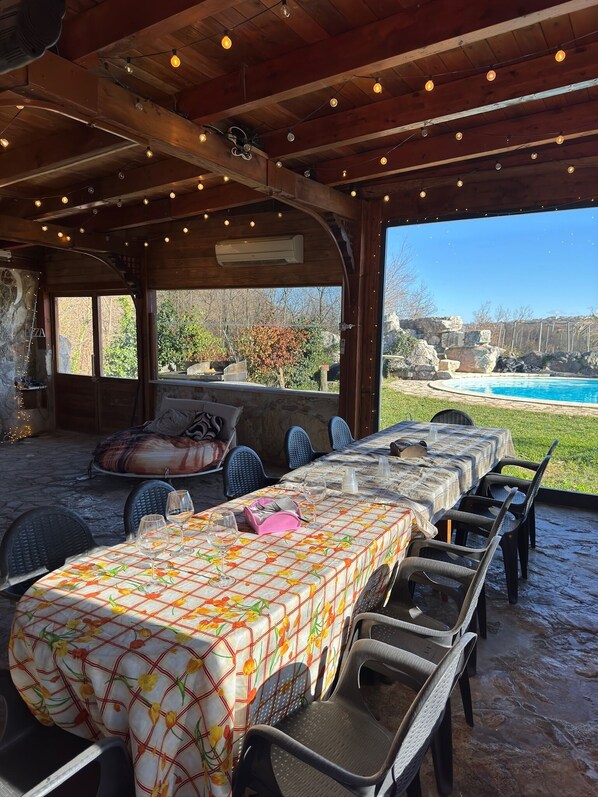 An outdoor patio with a long table set for dining, surrounded by chairs, and a view of a swimming pool in the background.