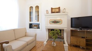 Een woonkamer met een beige bank, een houten salontafel, een televisie op een houten dressoir, een open haard met een schoorsteenmantel en een raam met een decoratieve omlijsting.