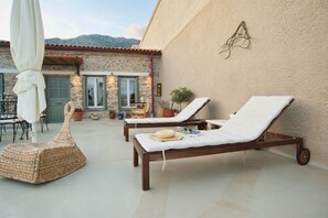 A sunlit outdoor lounge area with two wooden lounge chairs and a woven chair, a table with chairs, and a potted plant. - Guesthouse Simou with view (Arachova)