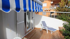 En balkon med en stribet markise, en hvid stol, liggestol og flisegulv.