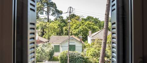 Janela com venezianas abertas, exibindo vista de um bairro residencial com casas, árvores e carros estacionados.