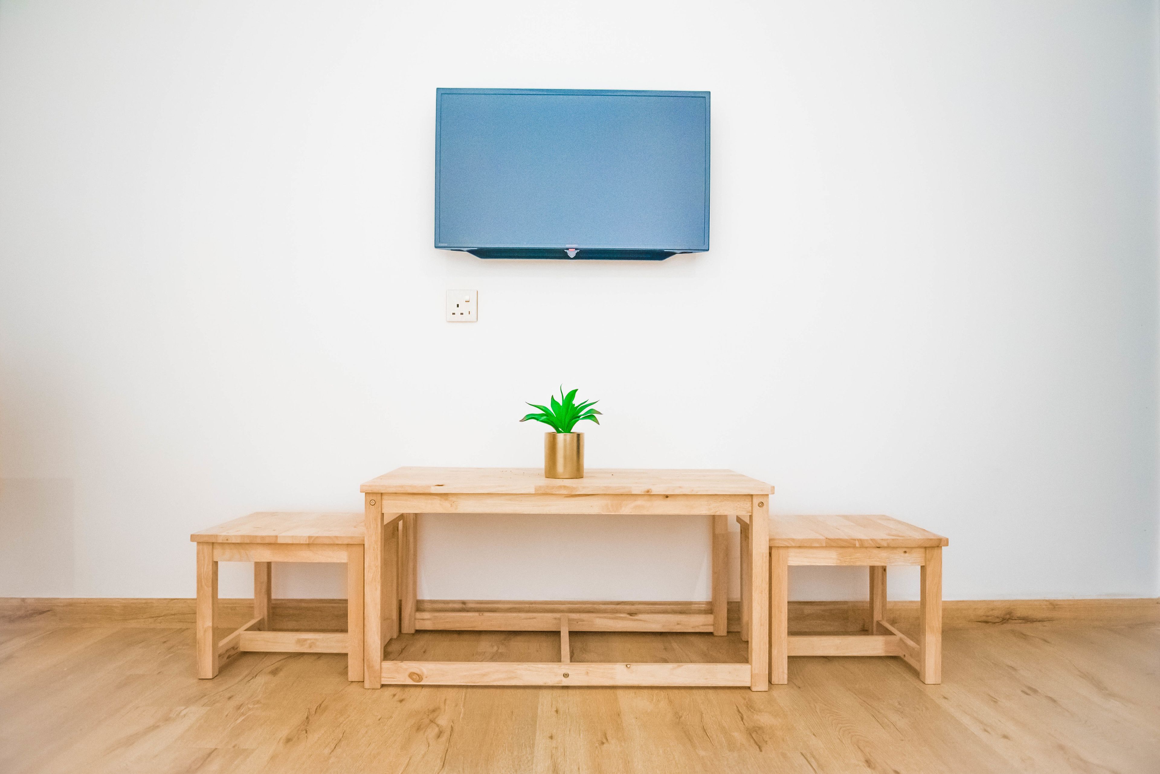 A room with a wooden table, two benches, a small potted plant, and a mounted television on the wall.