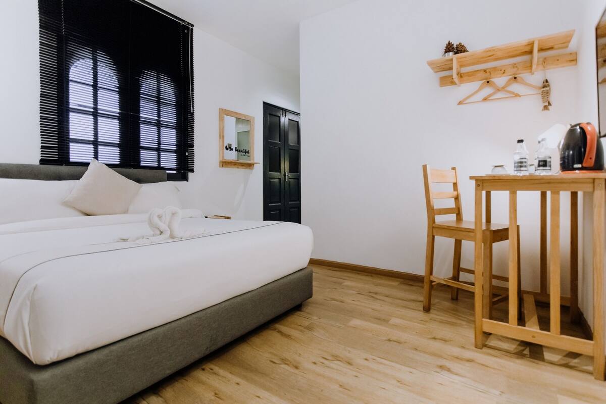 Ein Schlafzimmer mit einem Bett, einem Holztisch und einem Stuhl. Es gibt ein Fenster mit Jalousien, einen Spiegel und eine Tür.