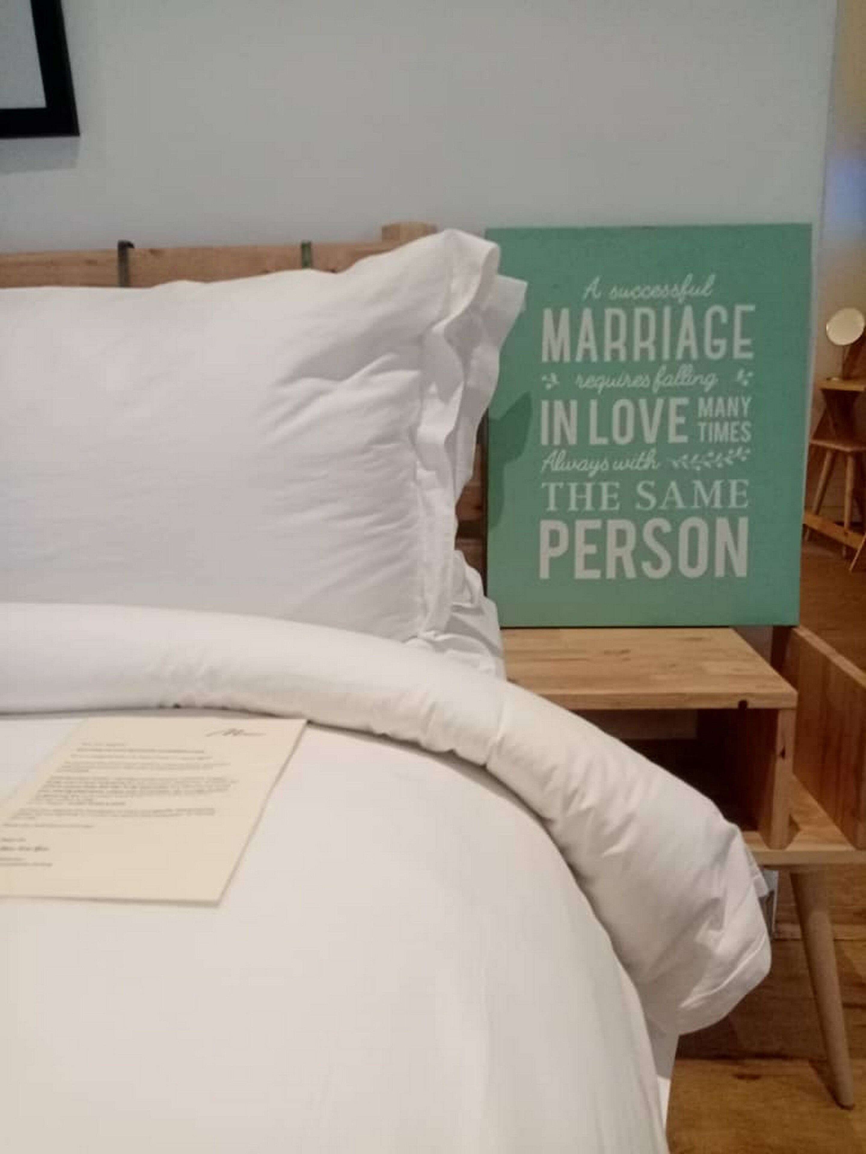 A neatly made bed with white linens, a wooden nightstand with a teal sign about marriage, and a framed picture on the wall.