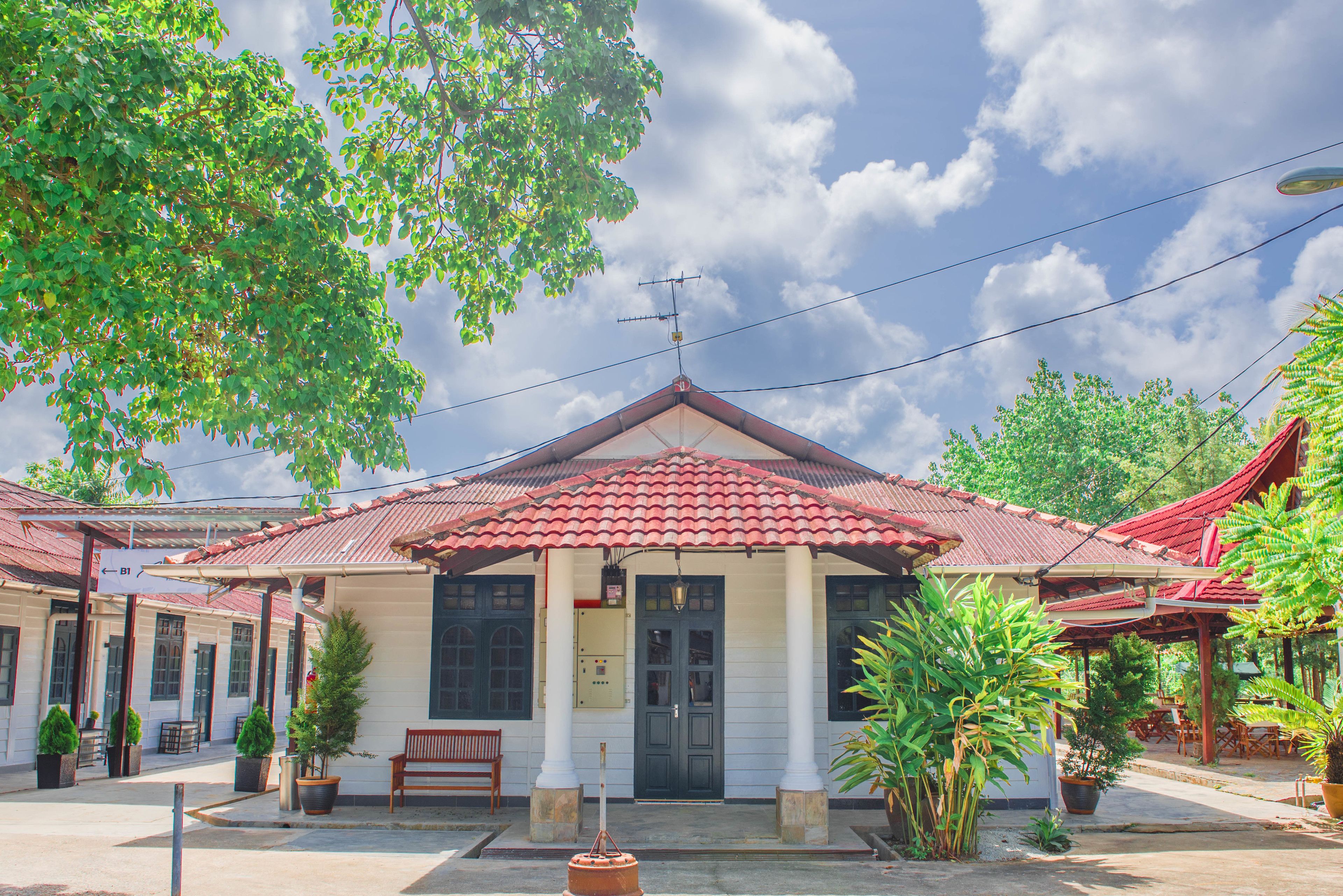 A building with a red-tiled roof, white walls, and a central entrance. There are potted plants and a bench outside.