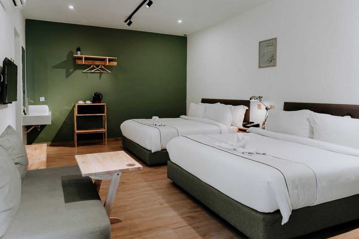 Ein Hotelzimmer mit zwei Betten, einem kleinen Tisch, einer Couch, einem Holzregal mit einem Wasserkocher und einem an der Wand montierten Fernseher.