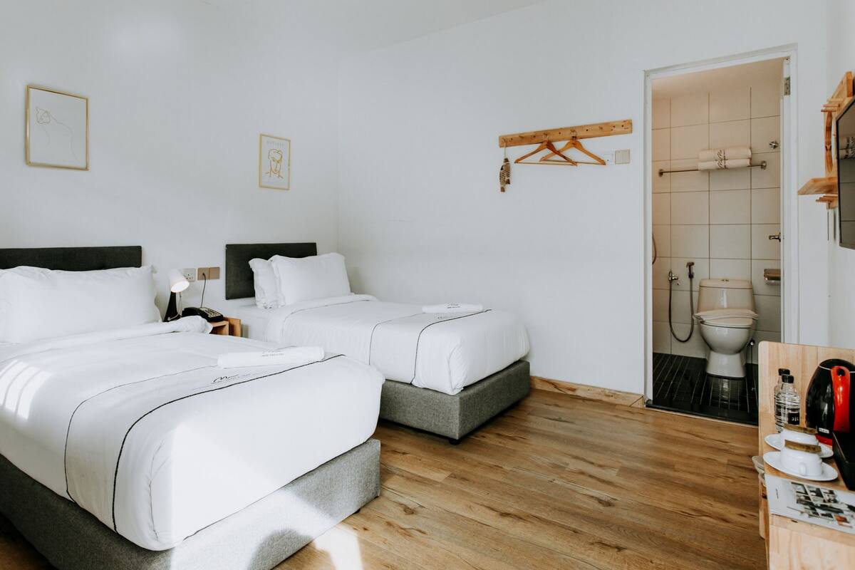 Ein Hotelzimmer mit zwei Betten, Holzboden, einem Badezimmer mit Toilette und Waschbecken sowie einem kleinen Tisch mit Wasserkocher und Tassen.
