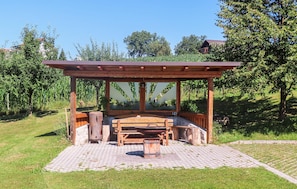 A wooden pergola with a bench and table, surrounded by a brick patio, set in a grassy area with trees and a house in the background. - Ferienhaus in Feldbach (Feldbach)