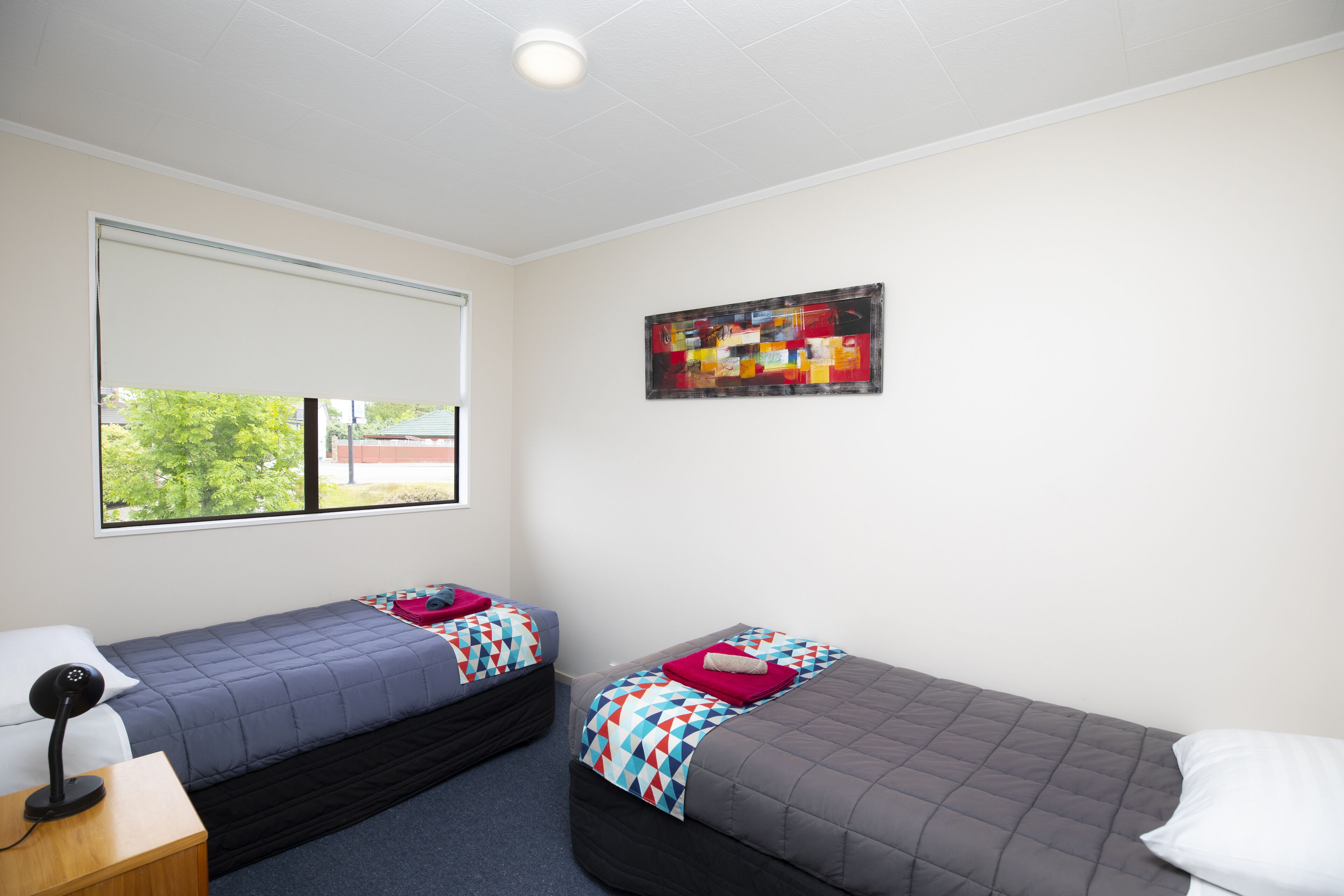 A room with two single beds, a desk with a lamp, a window with blinds, and a colorful abstract painting on the wall.
