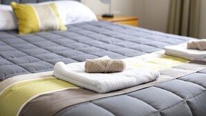 A neatly made bed with a grey quilt, a white pillow, and a yellow accent pillow. There is a small bedside table with a lamp and a folded towel on it. - Fitzgerald on King (Timaru)