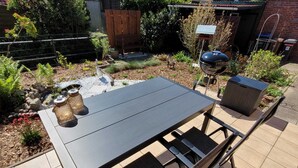 Eine Terrasse im Freien mit dunklem Tisch und Stühlen, umgeben von einem Garten mit einem kleinen Teich und verschiedenen Pflanzen.