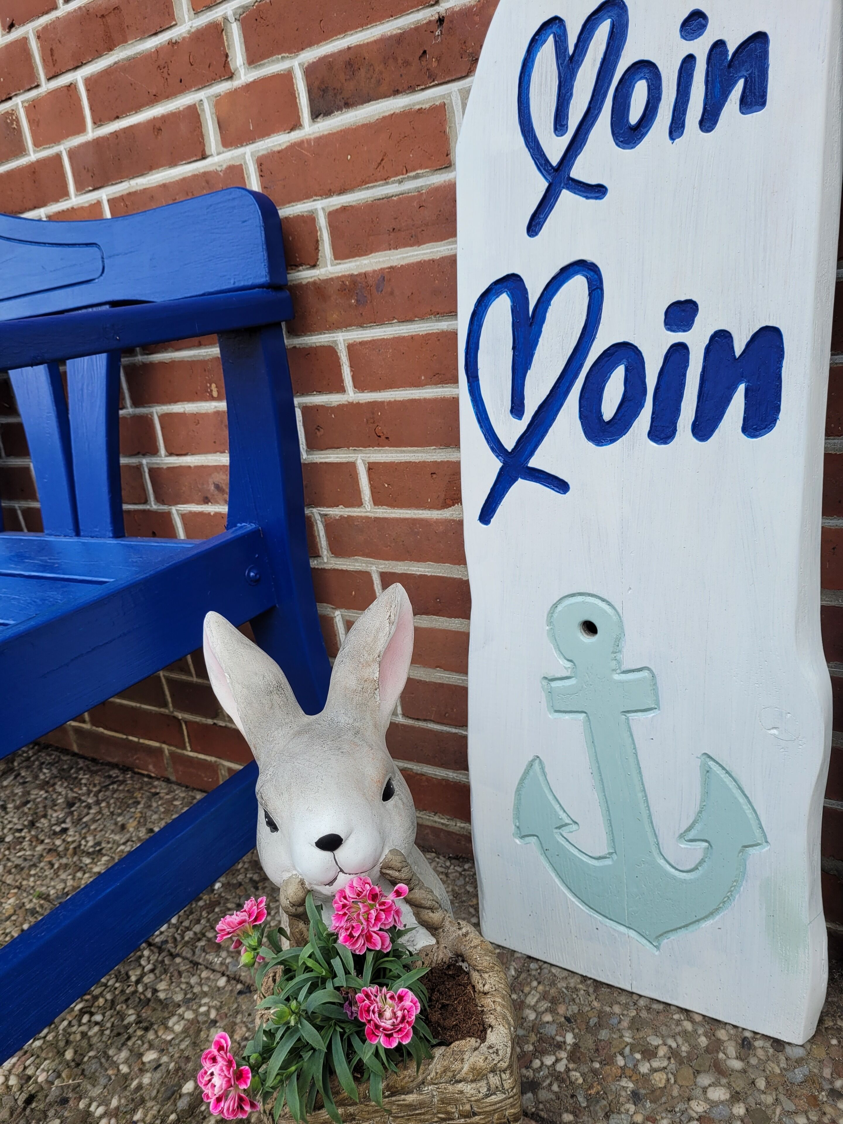 Ein weißes Schild mit blauer Schrift und einem Ankersymbol, platziert neben einer blauen Bank und einer Hasenfigur mit rosa Blumen.