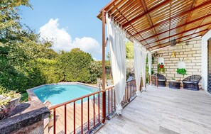 Un área de piscina con terraza de madera, una pérgola con techo de bambú y un muro de ladrillo con plantas en macetas.