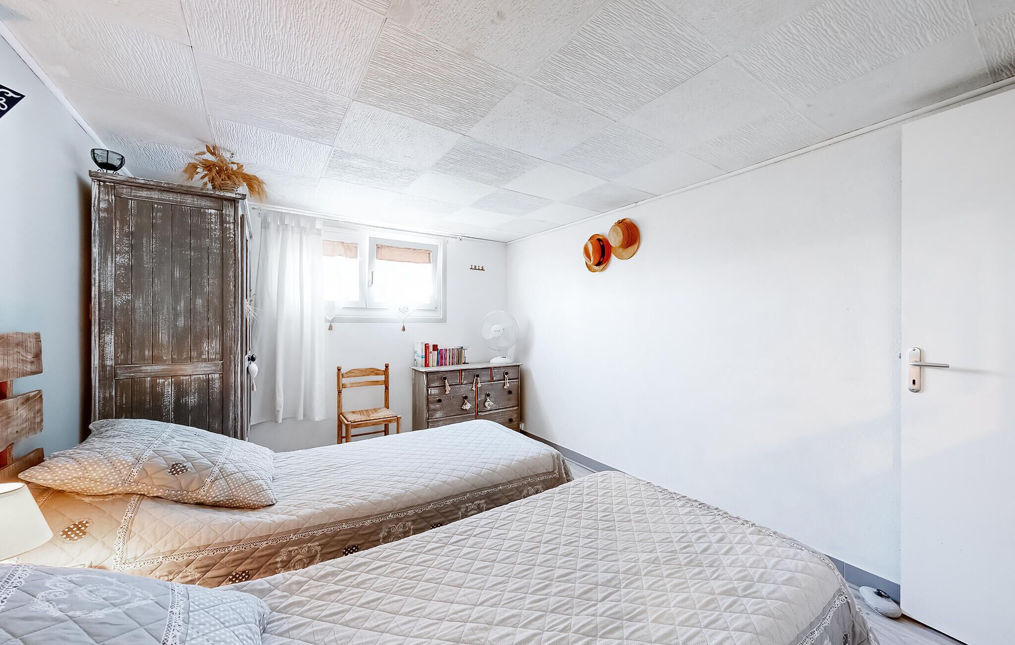 A bedroom with two beds, a wooden wardrobe, a desk with a chair, and a window with curtains.