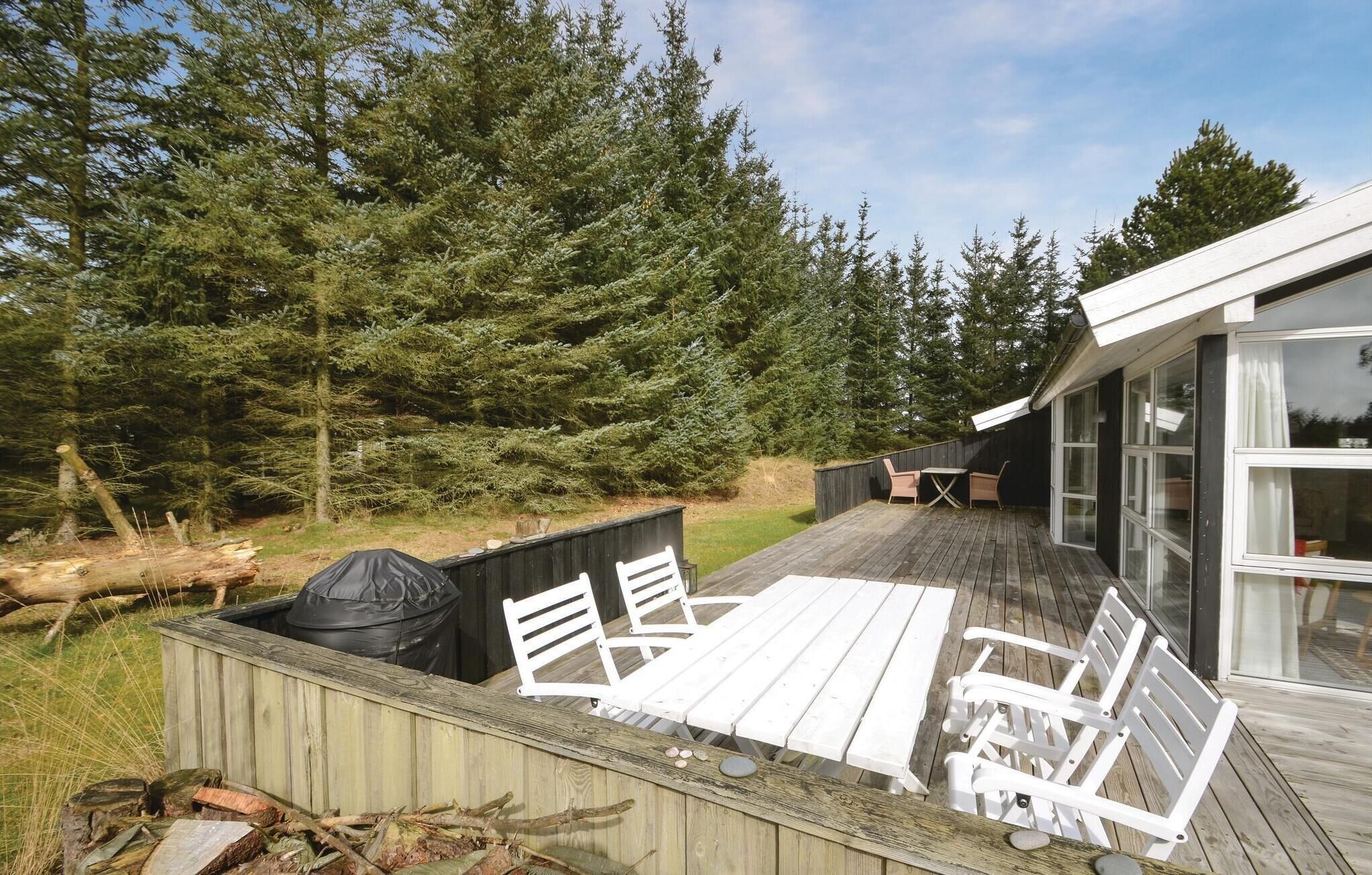 Una terraza de madera con mobiliario exterior blanco, un espacio acristalado y un fondo de bosque.