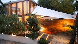 Een modern huis met grote glasramen, een houten constructie in de vorm van een boot en een tuin met planten en een pad.