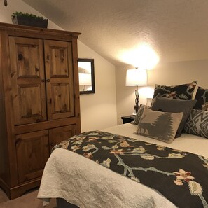 A bedroom with a large wooden wardrobe, a bed with a floral bedspread, and a nightstand with a lamp. - Charming Mountain Cottage. (Pine Valley)