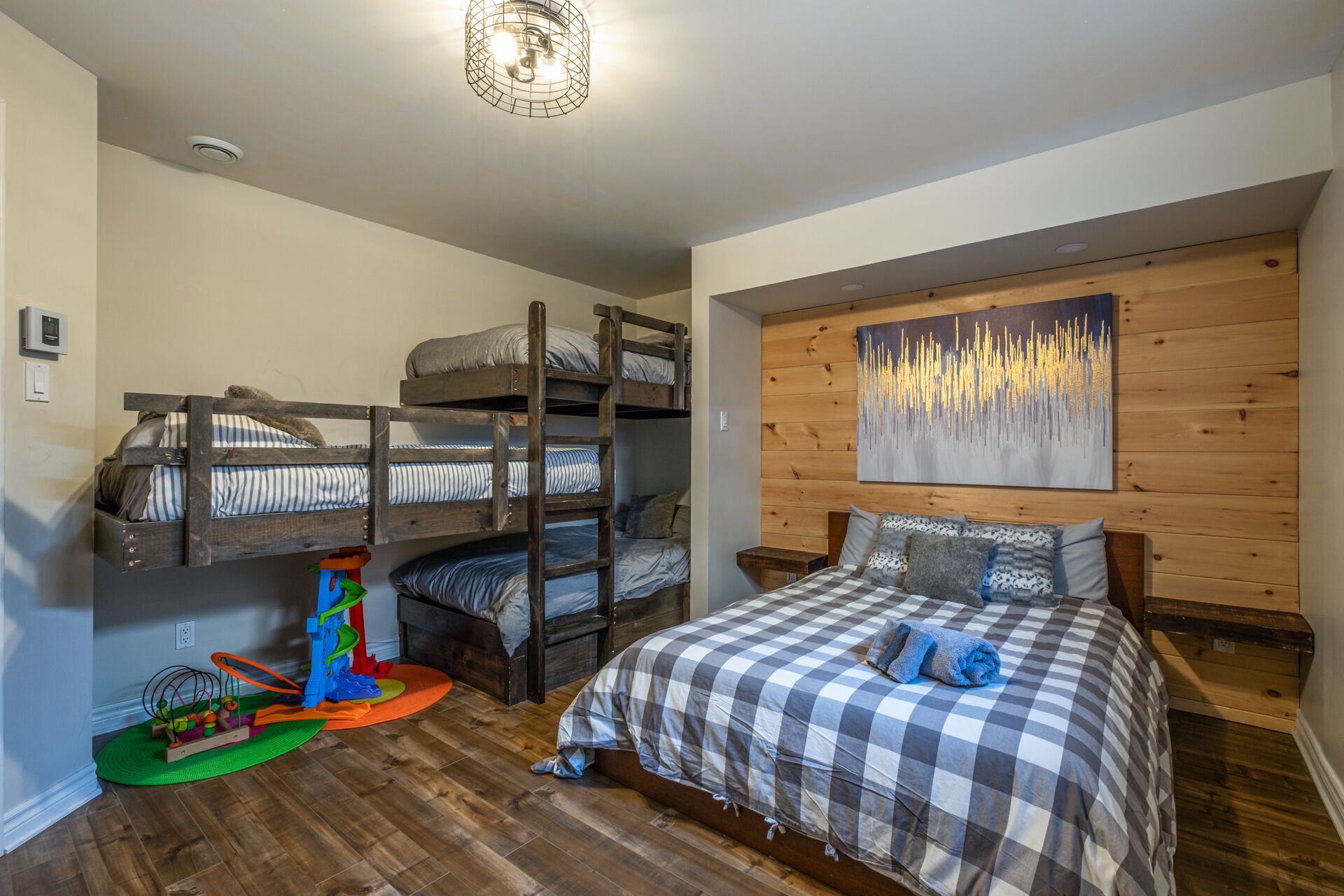Ein Zimmer mit Stockbetten, einem karierten Bett, einer Holzwand mit einem Gemälde und einer grünen Matte mit Spielzeug.