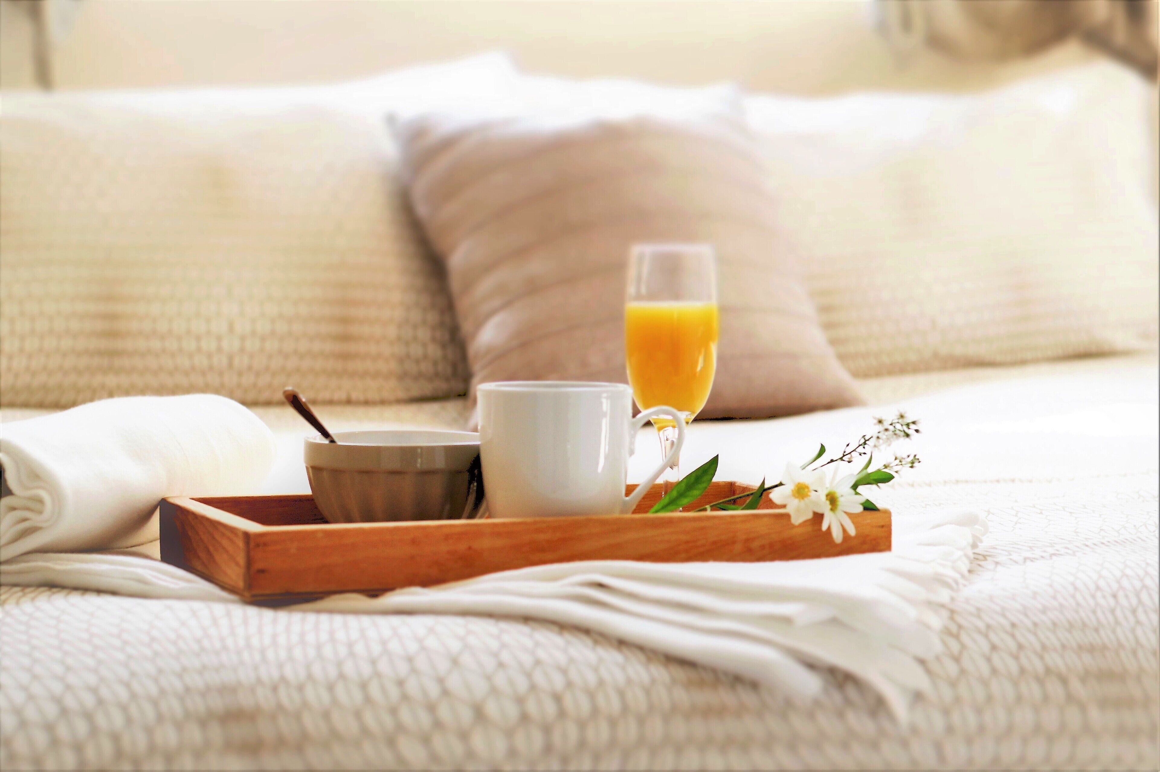 A wooden tray with a white mug, a glass of orange juice, and a bowl on a bed with white bedding and pillows.