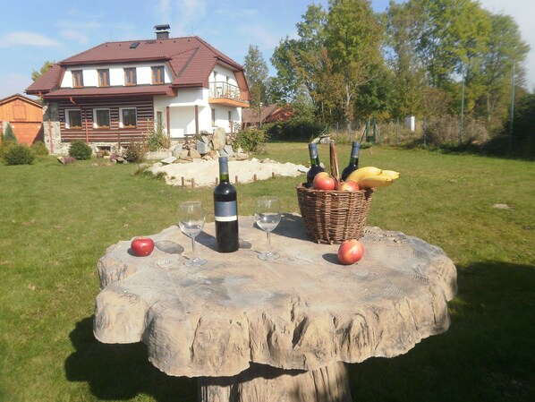 A picnic setup with a bottle of wine, glasses, and a basket of fruit on a wooden table in a grassy area with a house in the background.