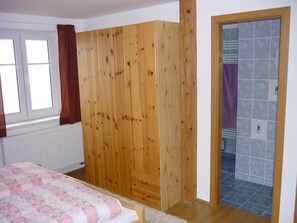A bedroom with a wooden wardrobe, a bed with a patterned bedspread, a window with curtains, and a bathroom with tiled walls.