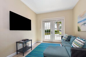 A living room with a sofa, a coffee table, a flat-screen TV mounted on the wall, and a sliding glass door leading to an outdoor area.