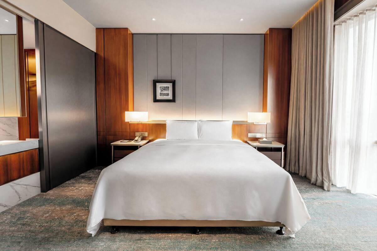 A hotel room with a large bed, two bedside tables, a nightstand, a framed picture on the wall, and a window with curtains.