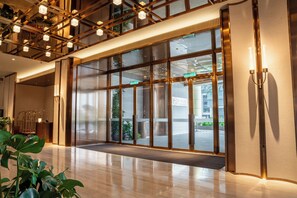 Un moderno vestíbulo de hotel con una amplia entrada de vidrio, suelo de madera y un techo con vigas a la vista y luces colgantes.