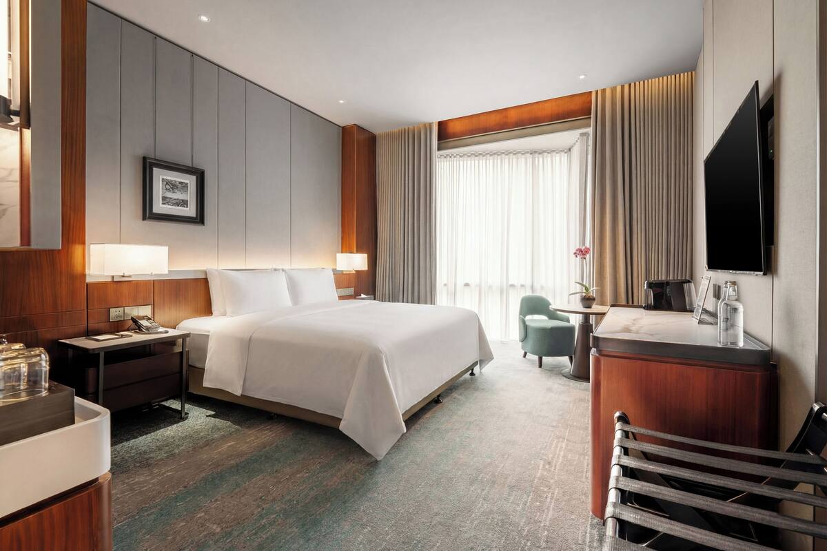 A hotel room with a large bed, bedside tables, a desk, a chair, a TV, and a window with curtains.