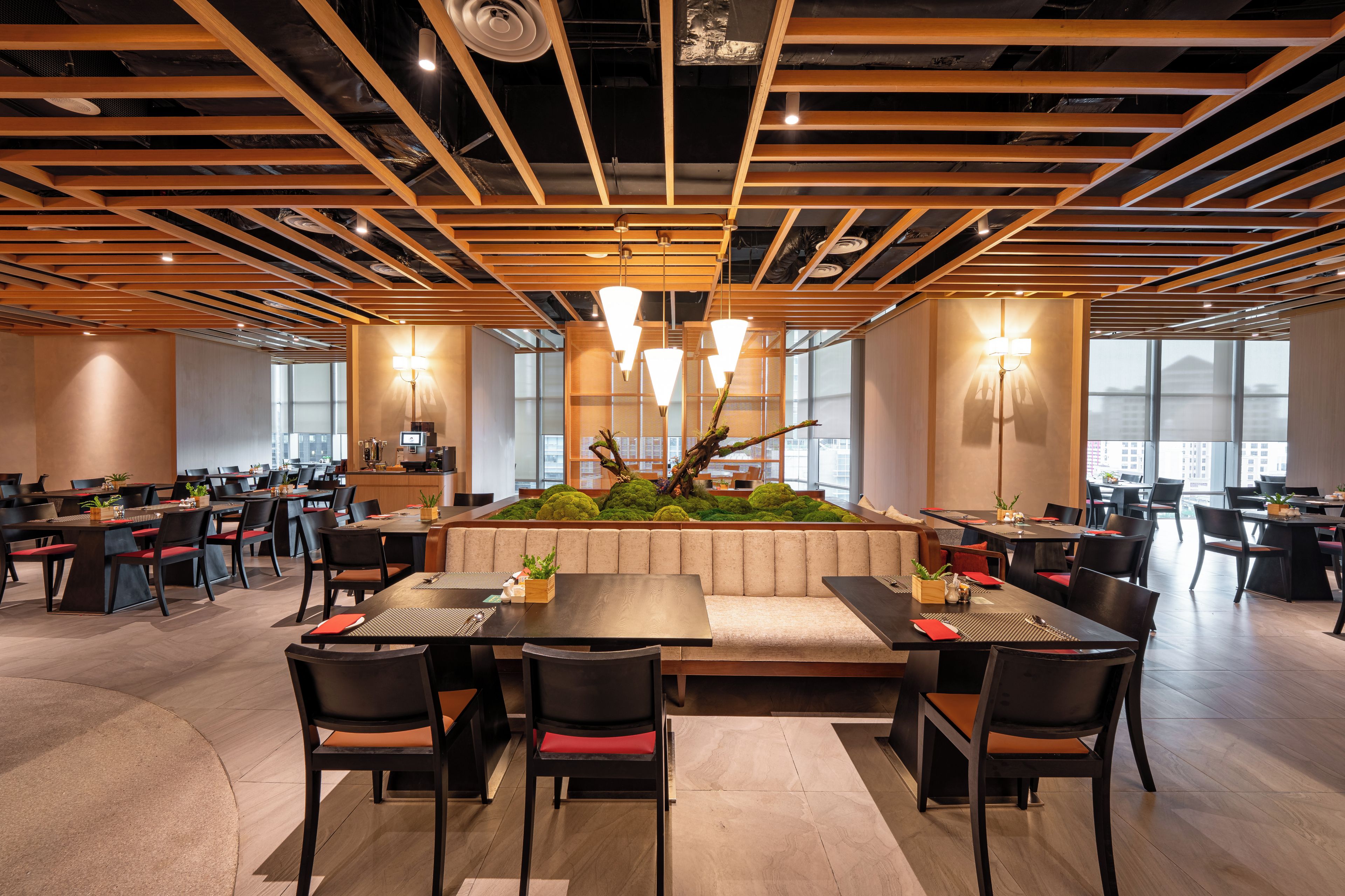 A spacious dining area with multiple tables and chairs, a central seating area with a decorative plant arrangement, and large windows providing natural light.