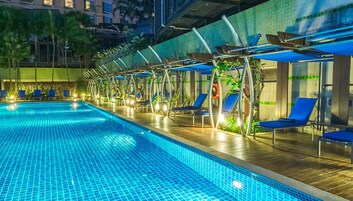 A swimming pool with blue tiles, surrounded by lounge chairs and umbrellas, with a wooden deck and a building in the background.