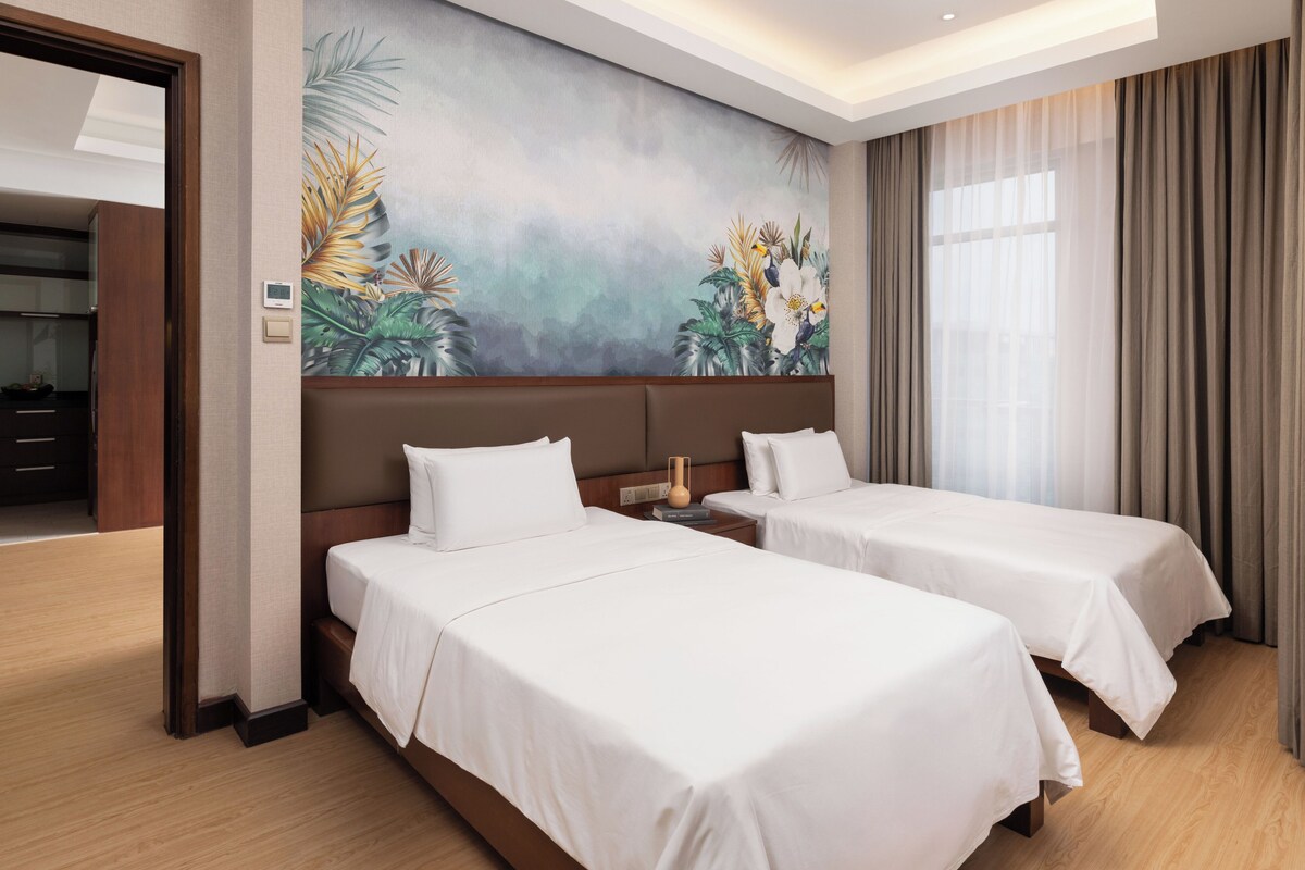 Habitación de hotel con dos camas, un mural grande en la pared, piso de madera y una ventana con cortinas.