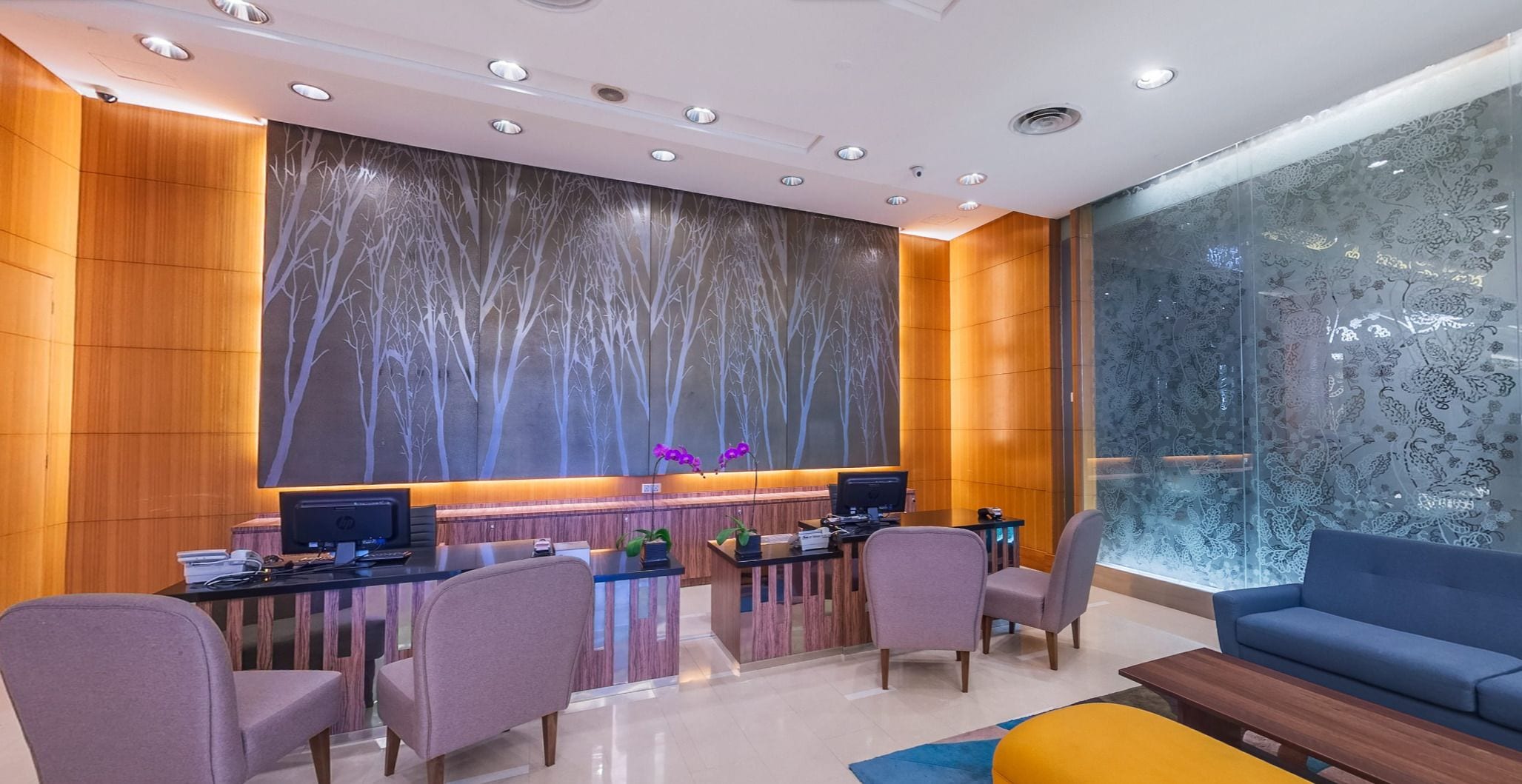 Una zona de recepción moderna con un panel de pared de madera, un ventanal con estampado floral y un área de descanso con sofá y sillones azules.