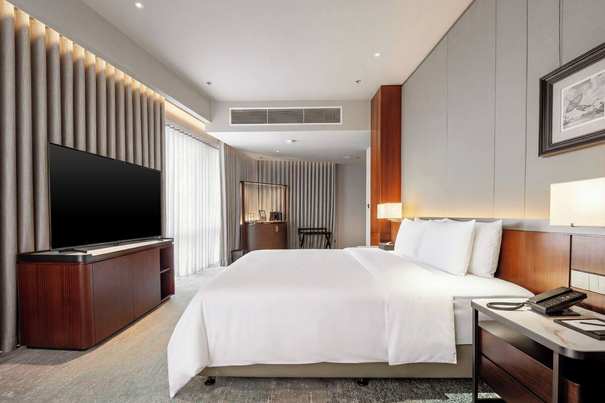 A hotel room with a large flat-screen TV, a bed with white bedding, a desk with a phone, and a lamp.