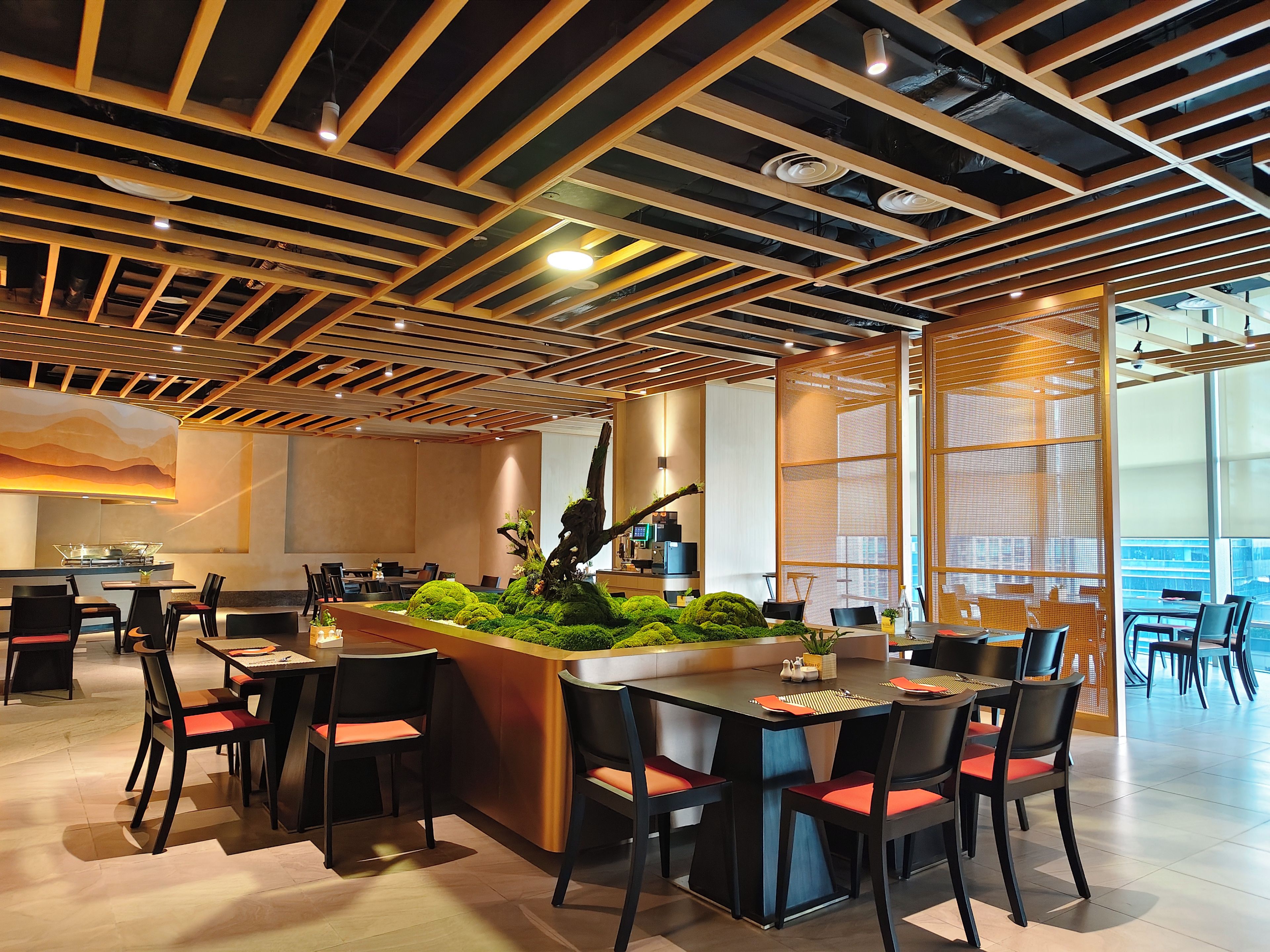 Een modern restaurantinterieur met houten balken in het plafond, een centraal eiland met beplanting en meerdere eettafels met stoelen.