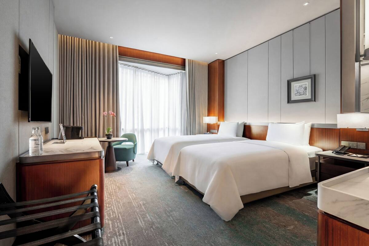 A hotel room with two beds, a desk, a chair, a TV, and a window with curtains.