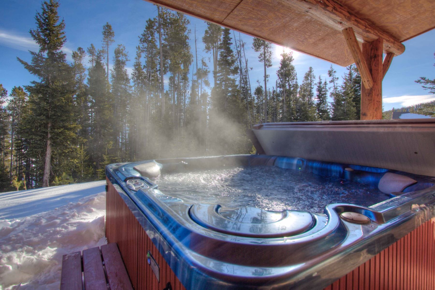 A hot tub with a wooden deck, surrounded by a snowy landscape and tall pine trees.