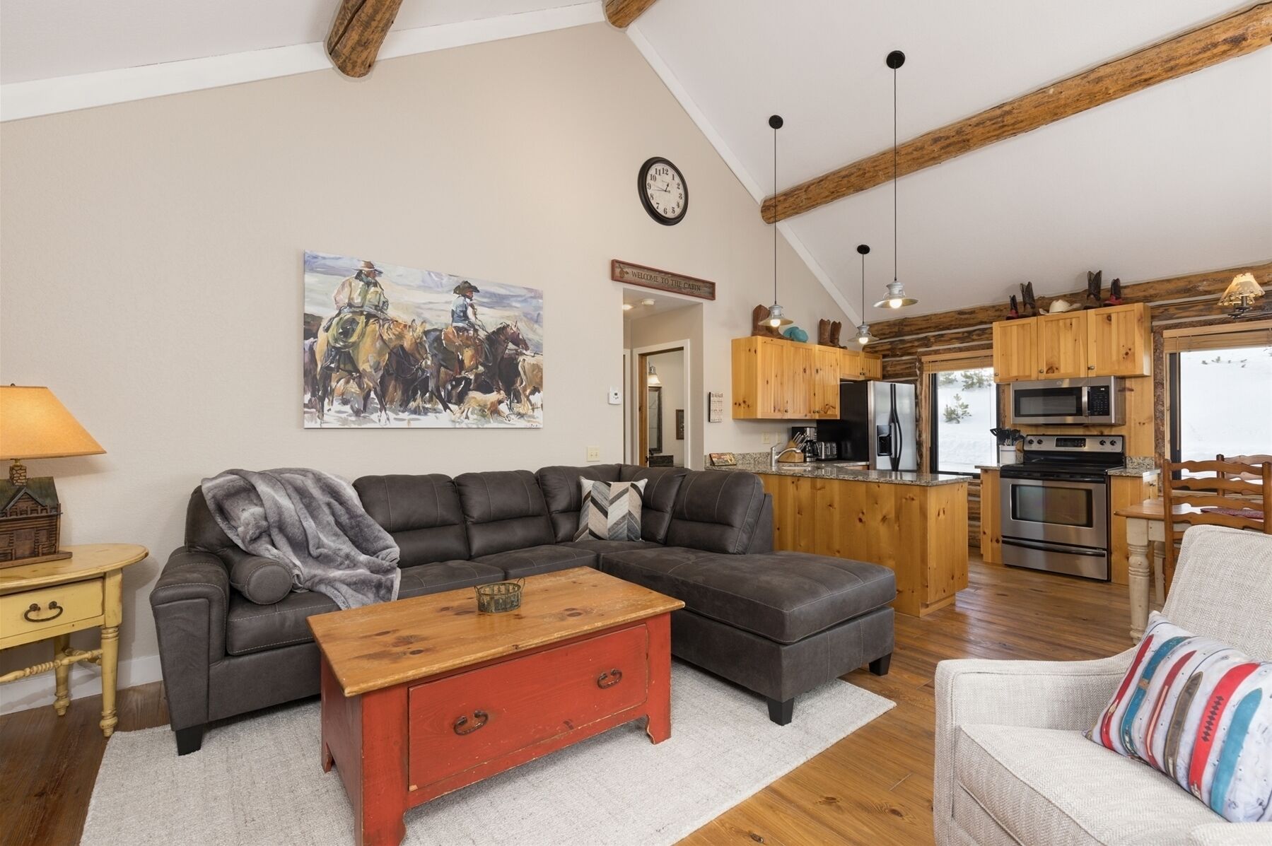 A spacious living room with a sectional sofa, a red coffee table, and a painting of cowboys on horseback. The kitchen features wooden cabinets and modern appliances.