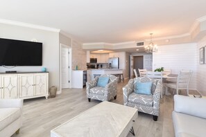 A modern living room with a flat-screen TV, patterned armchairs, a coffee table, and a dining area with a chandelier.