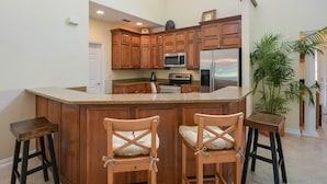 A kitchen with wooden cabinets, a stainless steel refrigerator, a microwave, and a large island with bar stools. - 3BR Golf Course Townhome | Community Pool, Full Kitchen & In-Unit Laundry (New Smyrna Beach)