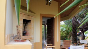 A covered outdoor seating area with wooden chairs and a table, a brick fireplace, and a door leading to the interior. - Situada dentro de um condomínio fechado de luxo, com a praia a 3 minutos a pé (Porto Santo)