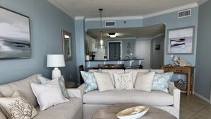 A living room with a beige sectional sofa, a round coffee table, and a dining area in the background.