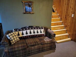 Un canapé à carreaux avec un plaid aux motifs de forêt, un petit tableau encadré accroché au mur et un escalier en bois.