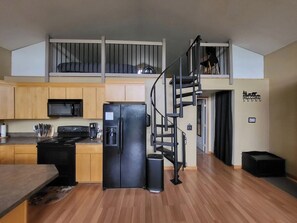 Eine moderne Küche mit einem schwarzen Kühlschrank, Herd und einer Mikrowelle. Ein Holztisch und ein schwarzer Mülleimer sind ebenfalls vorhanden. Eine Wendeltreppe verbindet die Küche mit einer oberen Ebene.