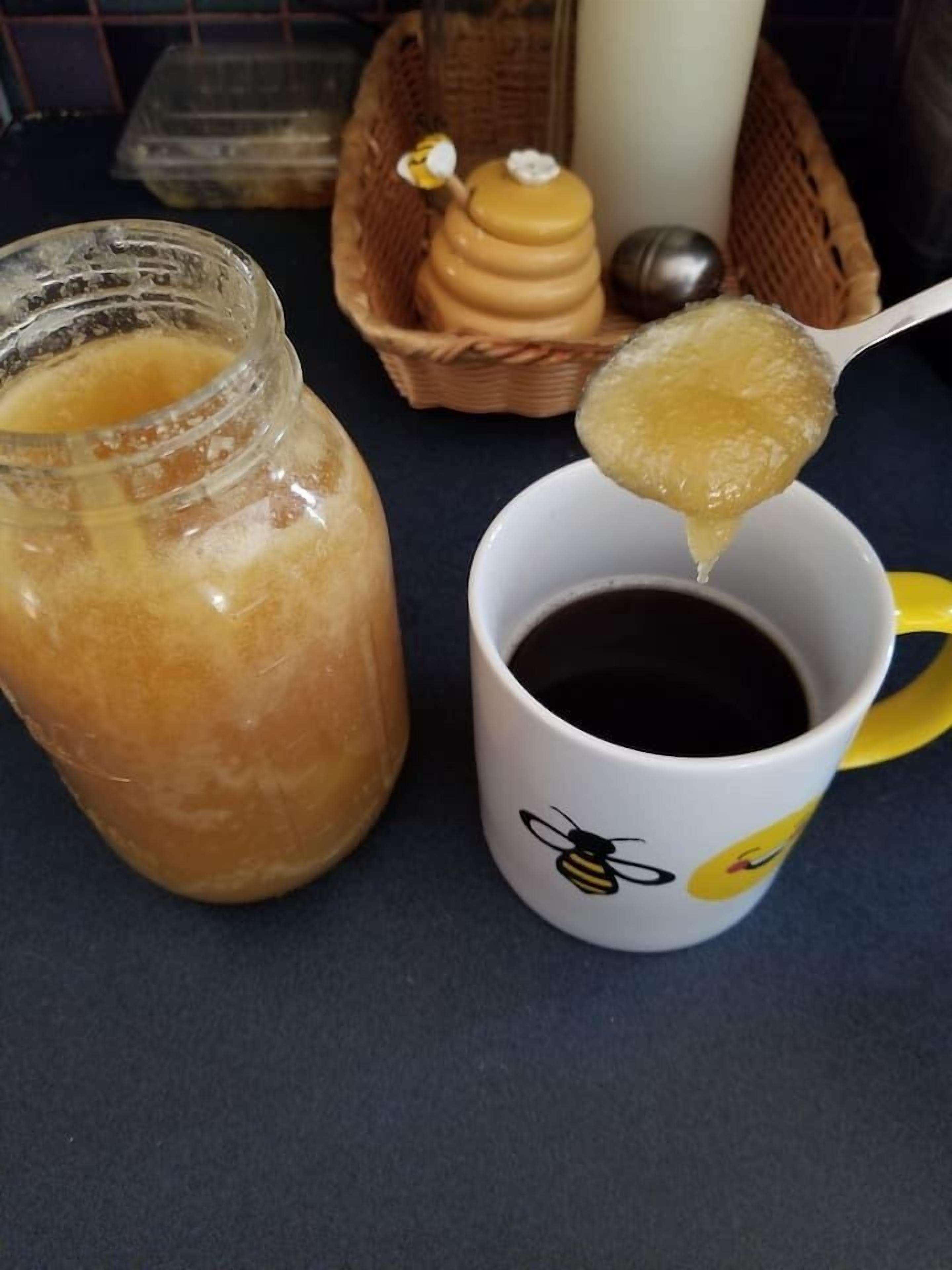 A jar of honey, a spoonful being poured into a mug, and a basket with honey jars and a candle in the background.