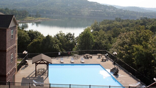 Piscina com espreguiçadeiras e um gazebo, cercada por árvores e vista para um lago.
