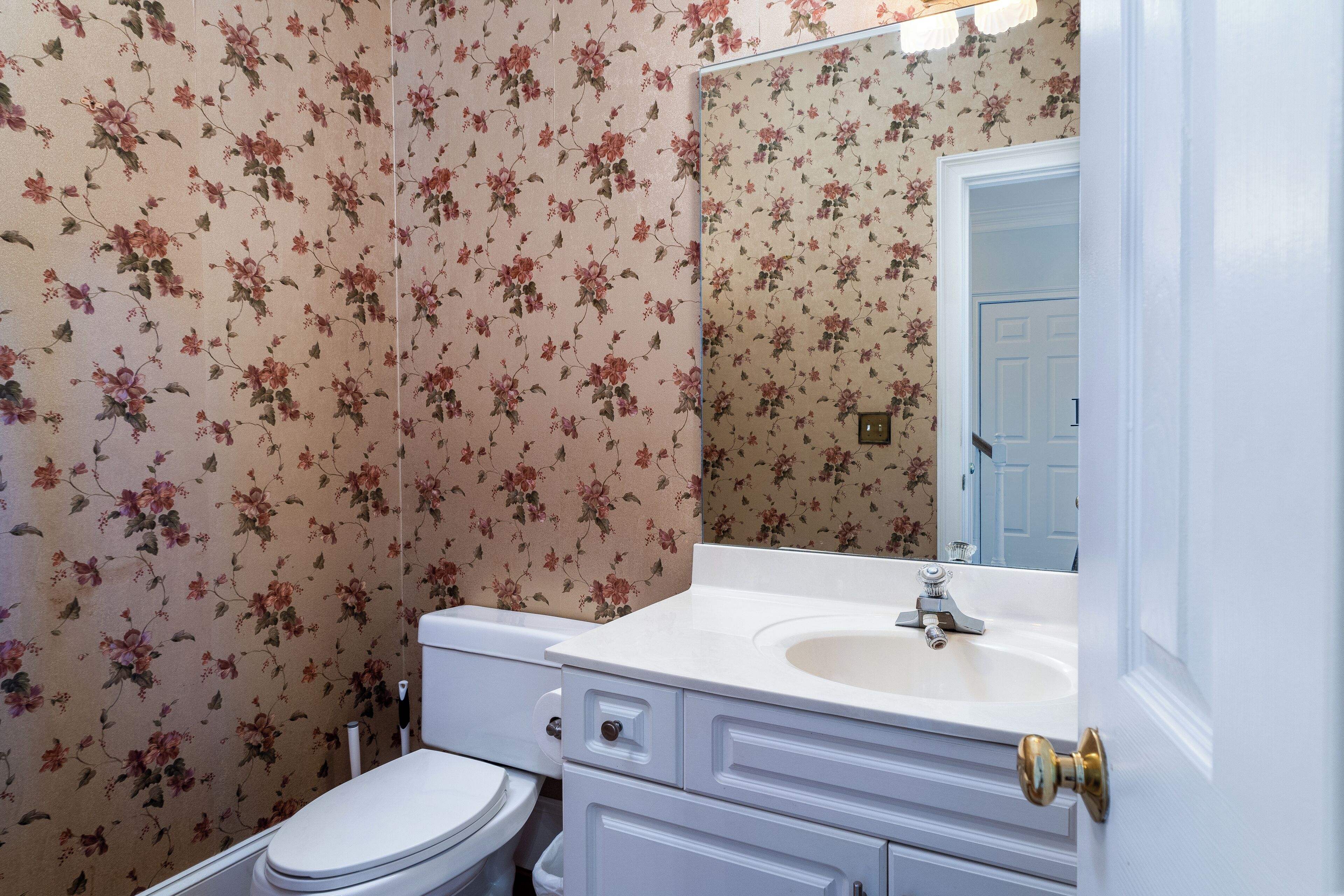 Une salle de bain avec un papier peint à motifs floraux, des toilettes blanches, un lavabo avec un miroir et une porte dotée d’une poignée en laiton.