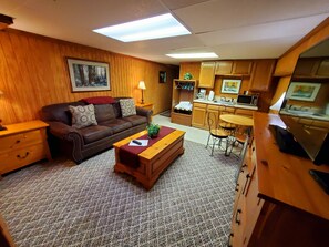 A cozy living room with a brown leather sofa, a wooden coffee table, and a kitchen area with wooden cabinets and a microwave.