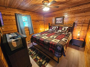 Ein gemütliches Schlafzimmer in einer Holzhütte mit Holzbalkendecke, einem Bett mit Patchworkdecke, einem Nachttisch, einer Lampe und einem Fenster mit Vorhängen.