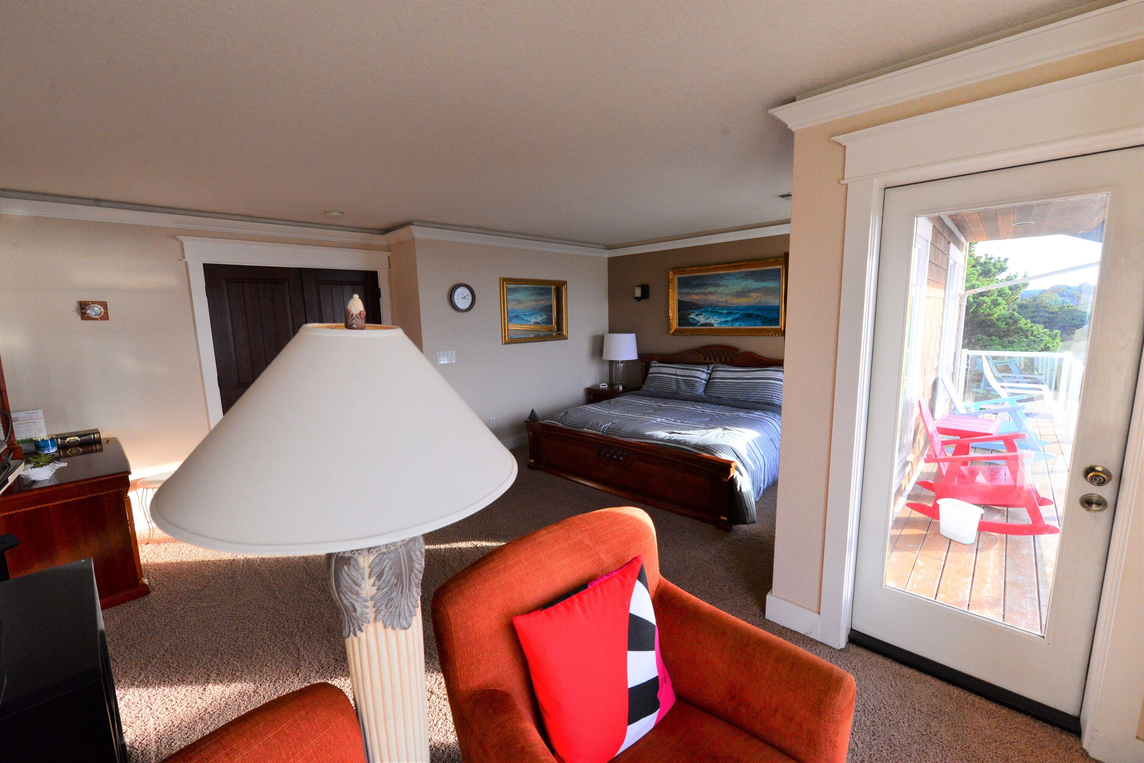 Ett hotellrum med en säng, en stol med en kudde, en lampa, en tavla på väggen och utsikt över den utomhusbelägna terrassen genom en dörr.